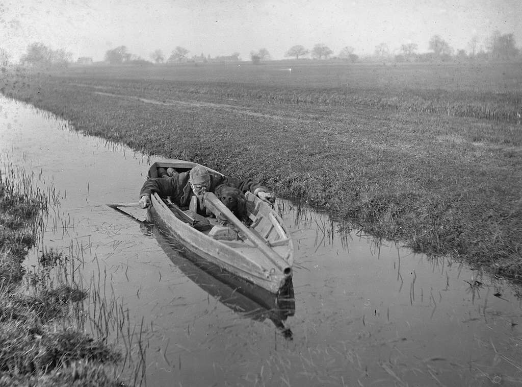 A man and a dog lay in a small boat on the water with a punt gun. 