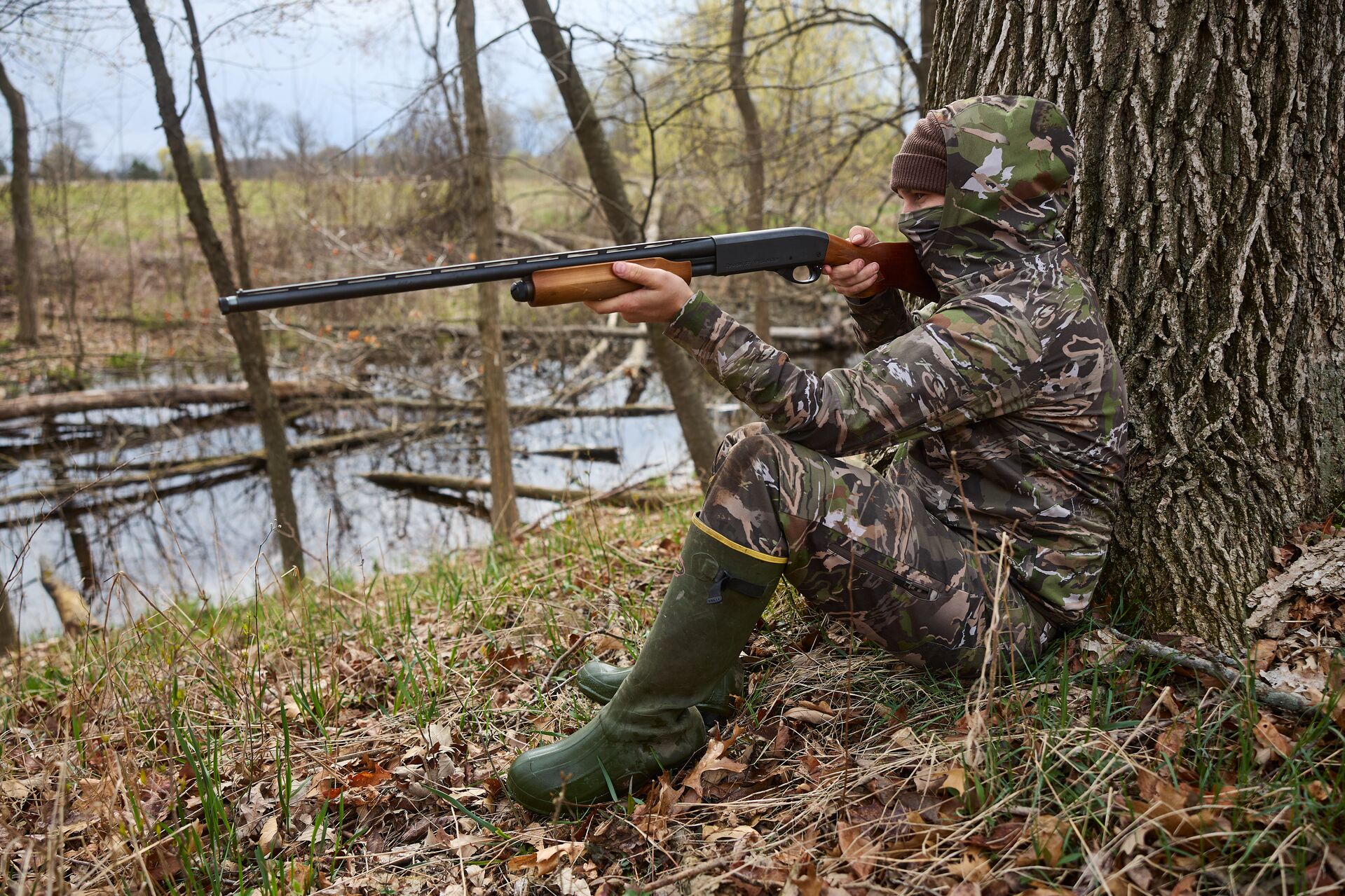 A hunter in camo sitting against a tree aiming a shotgun, hunting with a shotgun concept. 