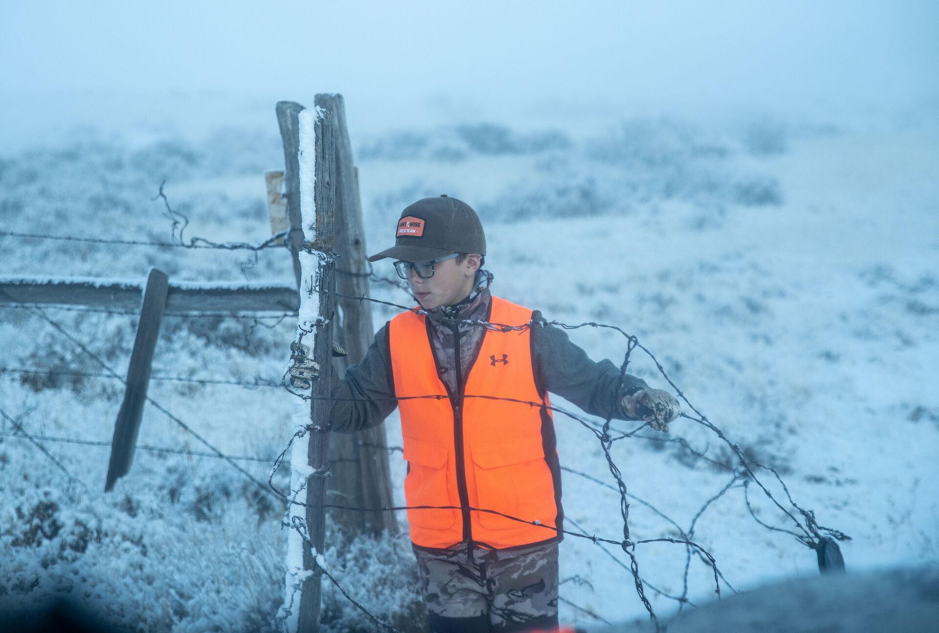 A young boy in blaze orange near a fence in the snow, ready to hunt. 