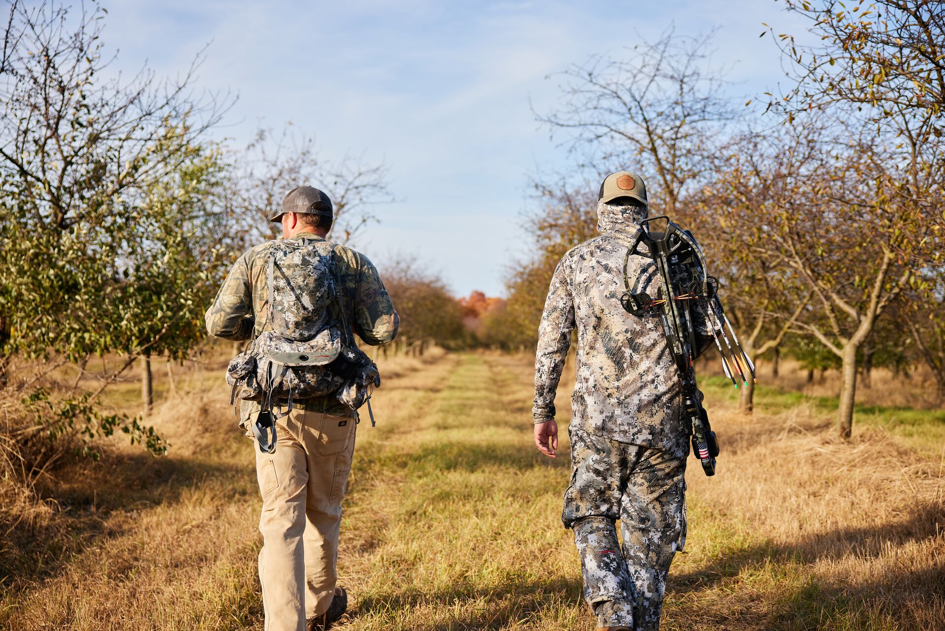 Two hunters dressed in camo walking in a field with hunting gear. 