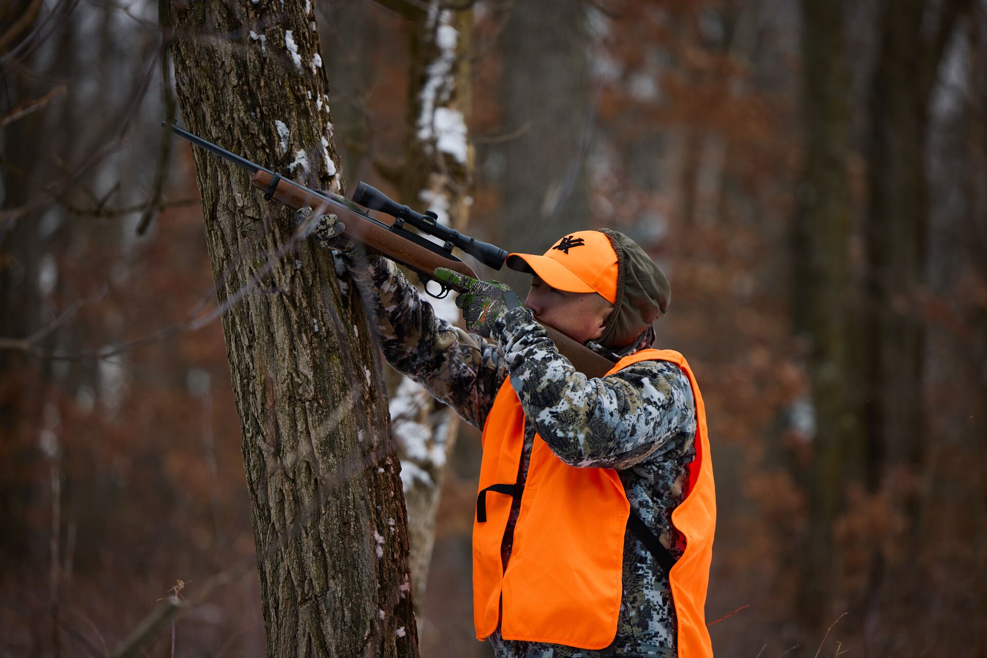 A hunter in blaze orange aims a rifle while in the woods. 