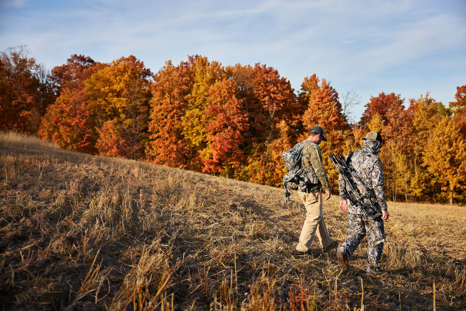 Two hunters hike across field with gear. 