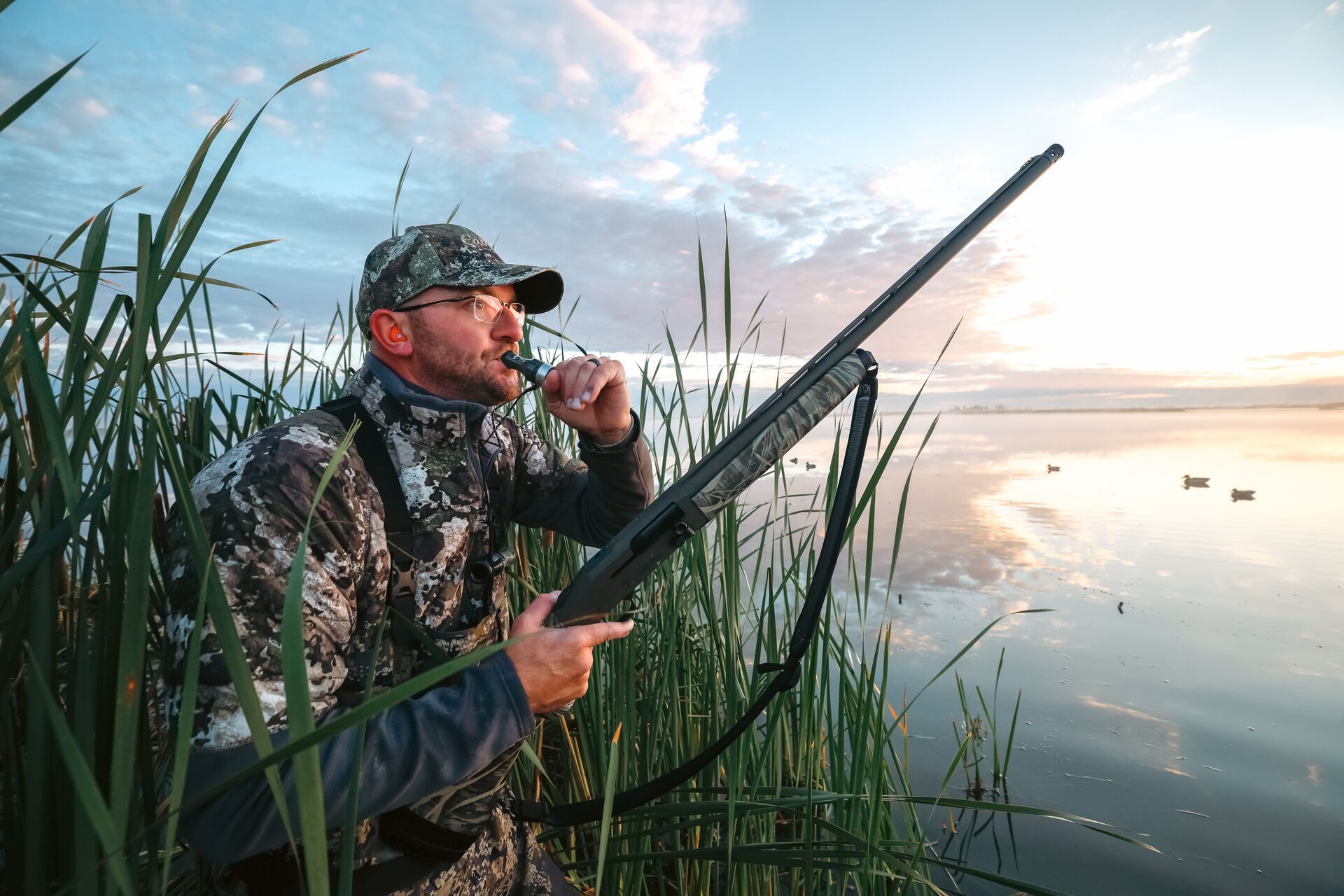 Duck hunter in tall reeds wearing camo with shotgun using duck call. 